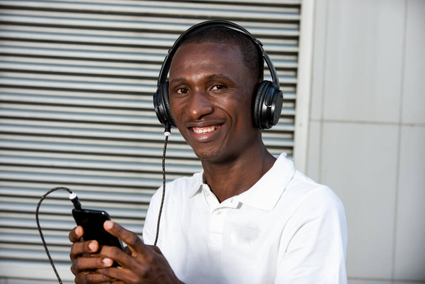 щасливий молодий чоловік у білій футболці слухає музику з гарнітурою мобільного телефону і співає на відкритому повітрі. Студент зі смартфоном та навушниками
 - Фото, зображення