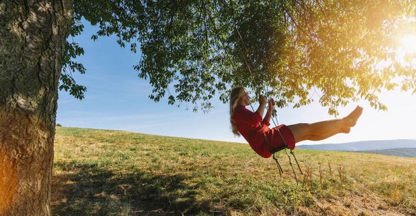 Cheerful lady in red sundress leaning back in her swing in sunny golden countryside. Evening sunshine beaming on lovely girl swaying under single tree in big green meadow - Photo, Image