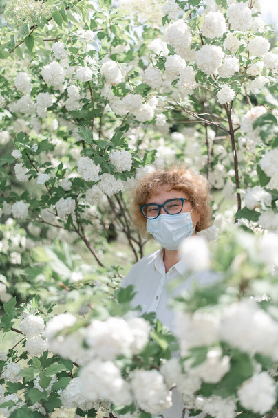 An elderly woman in glasses and a medical mask among the flowering bushes of an apple tree during quarantine. Spreads of coronavirus in spring. Grandmother walks in the park during isolation. - Fotoğraf, Görsel