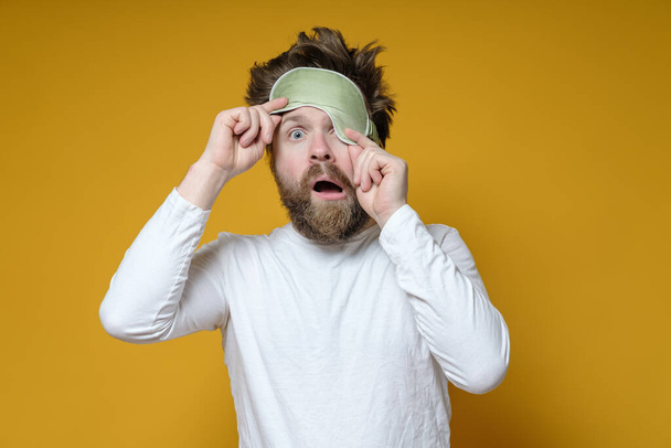Mann mit zerzauster Frisur hob Schlafmaske von einem Auge und sieht mit offenem Mund schockiert aus. - Foto, Bild