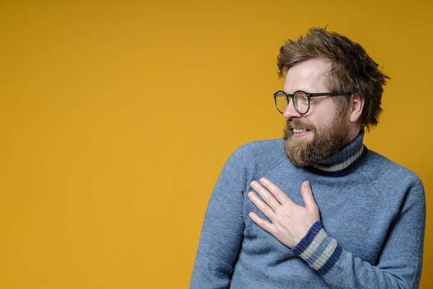 Cute modest man holds his hand on his chest and smiles good-naturedly. Friendly hipster in an old sweater. Copy space. - Photo, Image