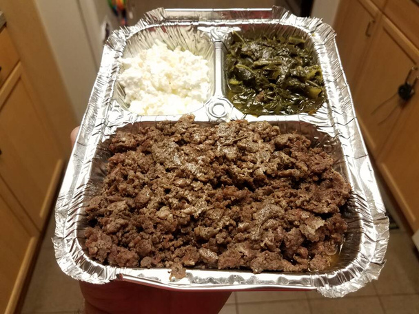 hand holding Ethiopian food beef and greens and cheese on metal tray in kitchen - Photo, Image