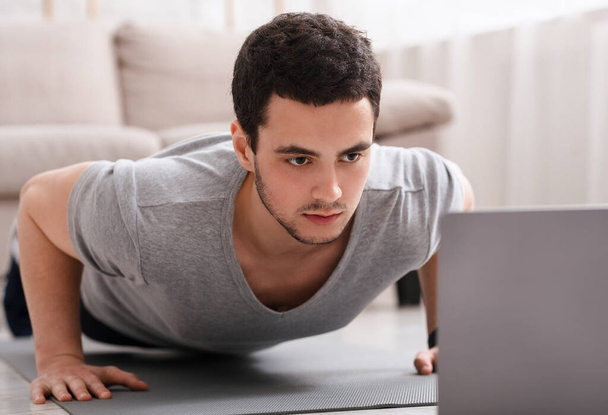 Fernarbeit und Online-Sport. Kerl ist Liegestütze und schaut auf Laptop im Innenraum - Foto, Bild
