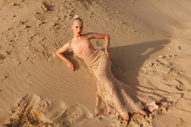 Blonde woman in splendid gold dress posing in desert, lying on the sand at the sunset. Horizontal view. - Fotografie, Obrázek