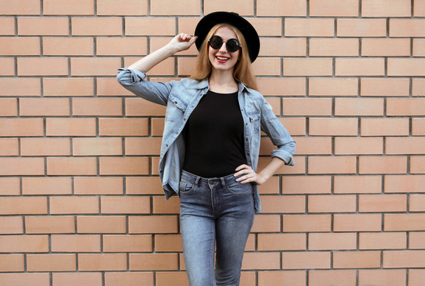 Happy smiling young woman wearing a black round hat, casual jeans jacket, female model posing on brick wall background - Zdjęcie, obraz