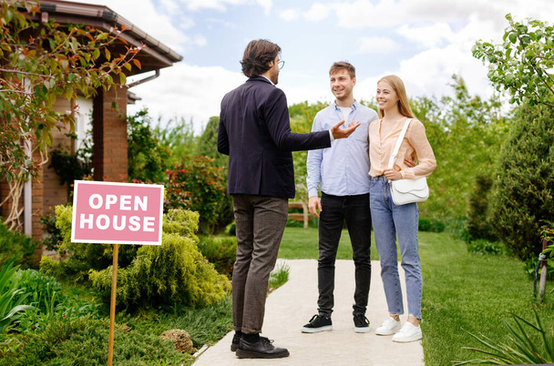 Confident realtor showing house for sale to newlywed young couple, outdoors - Photo, Image