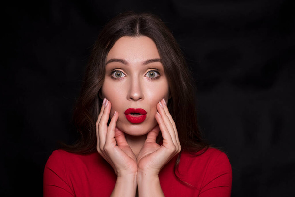 Emotional head shot portrait of a brunette caucasian woman in red dress and with red lips on black background. She make surprise, astonishment emotions with hands near her face. - Photo, Image