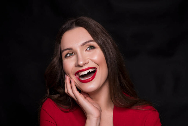 Emotional head shot portrait of a brunette caucasian woman in red dress and with red lips on black background. She smiling with hands near her face. - Photo, Image