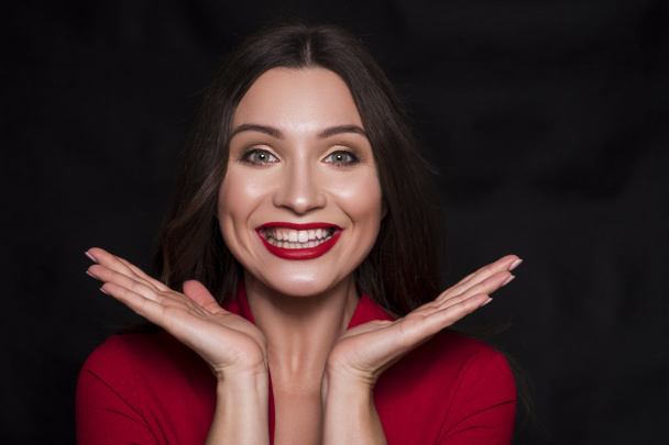 Emotional head shot portrait of a brunette caucasian woman in red dress and with red lips on black background. She look happy, smiling, with hands near her face. - Φωτογραφία, εικόνα