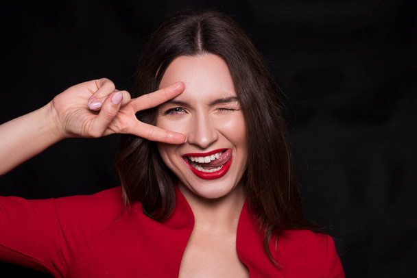 Emotional head shot portrait of a brunette caucasian woman in red dress and with red lips on black background. She make funny emotion with fingers near her eye and licking lips with tongue. - Photo, Image