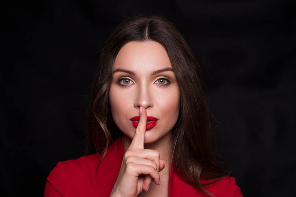 Emotional head shot portrait of a brunette caucasian woman in red dress and with red lips on black background. She make Shh emotion with finger near her lips. - Photo, Image