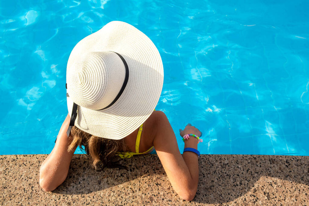 Vista dall'alto verso il basso di una giovane donna che indossa un cappello di paglia gialla che riposa in piscina con acqua cristallina blu nella giornata estiva di sole. - Foto, immagini