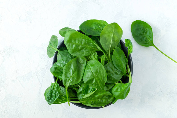 Fresh spinach leaves in a black bowl on a light surface. Fresh greens. Vegetarianism, healthy eating concept. Horizontal orientation, selective focus. View from above. - Foto, afbeelding