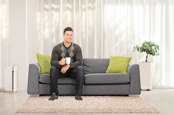 Full length portrait of a casual guy sitting on a sofa at home and holding a cup of tea - Photo, image