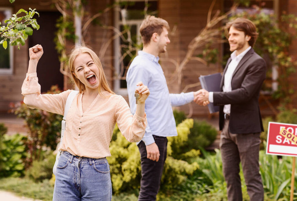 Ecstatic millennial woman with house key and her boyfriend shaking hands with real estate broker in backyard - Foto, Bild