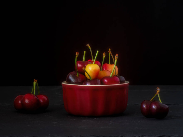 Yellow and red cherries in a red container against black background  artistic still life with healthy food with copyspace.  - Foto, imagen