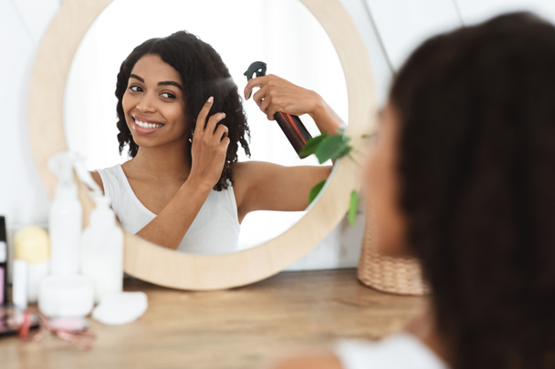 Penteado. Mulher negra bonita usando spray de cabelo para fácil pentear perto do espelho
 - Foto, Imagem