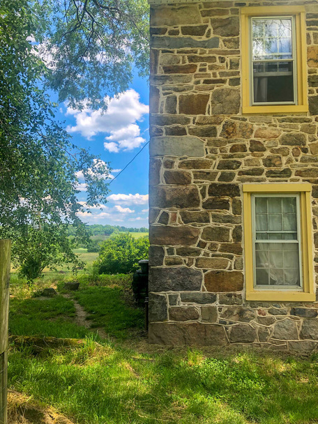 Old fieldstone masonry farmhouse sits on soft grass, green farm and blue sky background and dappled sunlight. Soft and rough textures, historic hefty stone construction yellow windows, copy space. - Photo, Image