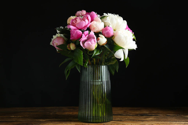 Bouquet of beautiful peonies in vase on wooden table against black background - Photo, image