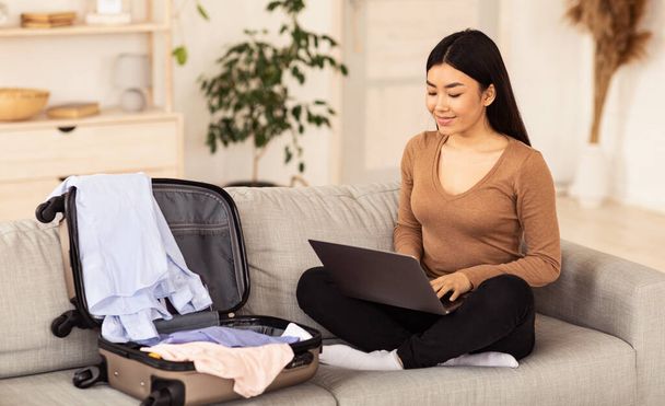 Chinese Girl Using Laptop Sitting With Unpacked Suitcase At Home - Foto, Imagem