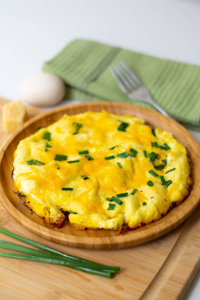 Fluffy omelette from whipped eggs with cheddar cheese and green onion scallions sprinkled on it in a wooden plate. It is surrounded by scallions, an egg, basil leaves, cheese, a green napkin, a fork - Photo, Image