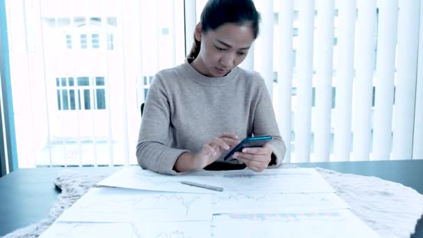 Uma mulher asiática segurando um smartphone deixa seu rosto triste. Desapontado de fazer negócios Há um gráfico de ações na mesa
. - Filmagem, Vídeo