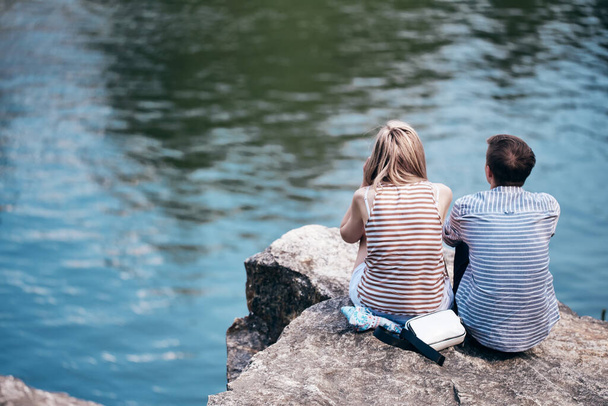 男と女の子は川の上の崖の上に座って前を見ている。リアビューだ。恋人同士だ。縦の写真。美しい自然と青い水. - 写真・画像