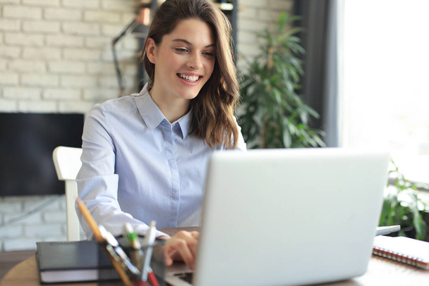 Smiling pretty woman sitting at table, looking at laptop screen. Happy entrepreneur reading message email with good news, chatting with clients online - Foto, Imagen