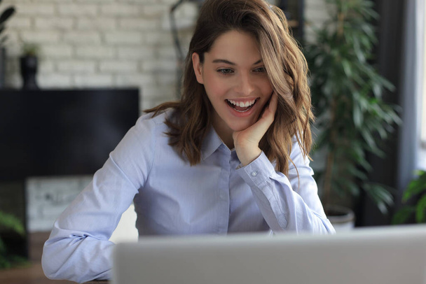 Smiling pretty woman sitting at table, looking at laptop screen. Happy entrepreneur reading message email with good news, chatting with clients online - Foto, Imagem