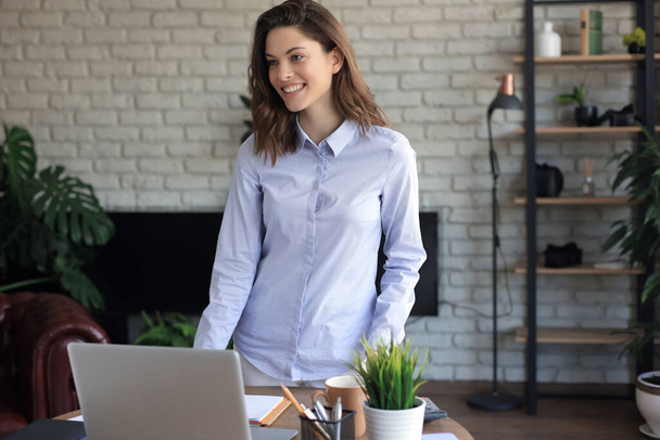 Confident young businesswoman with a friendly smile standing behind her desk in a home office - Photo, Image