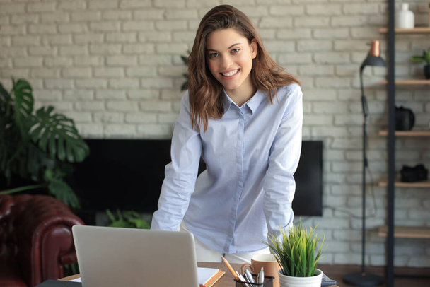 Confident young businesswoman with a friendly smile standing behind her desk in a home office looking at the camera - Photo, image