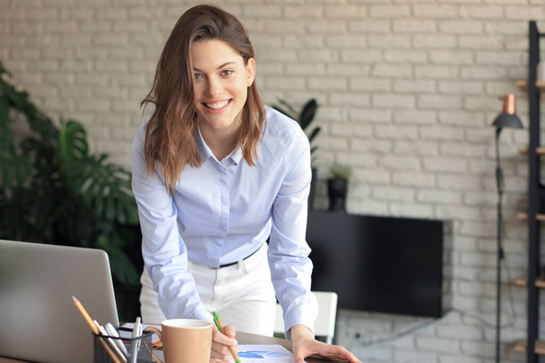 Confident young businesswoman with a friendly smile standing behind her desk in a home office looking at the camera - Photo, image