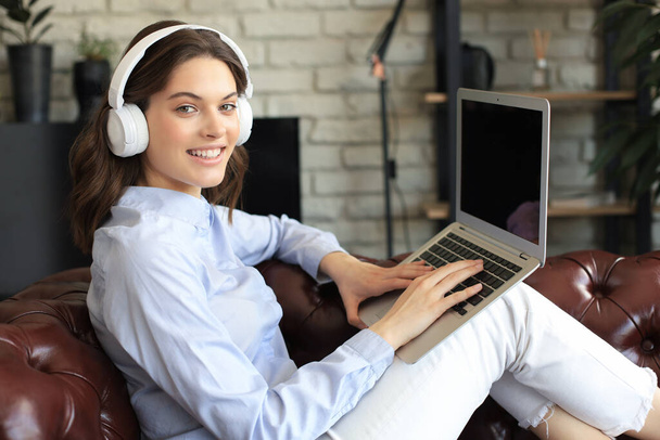 Smiling young woman with headphones and laptop on the sofa - 写真・画像