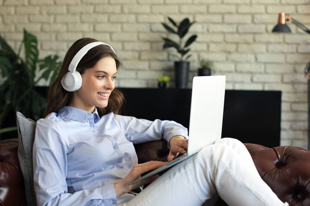 Smiling young woman with headphones and laptop on the sofa - Fotó, kép