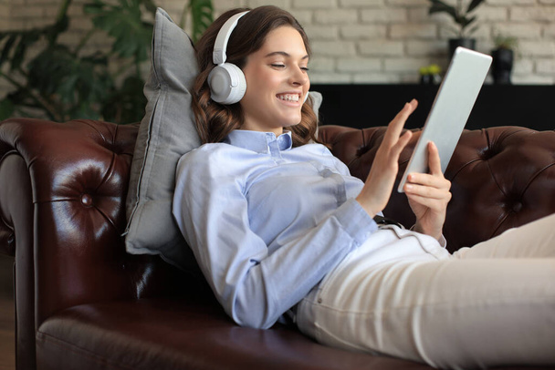 Smiling young woman with headphones and digital tablet on the sofa - Foto, afbeelding