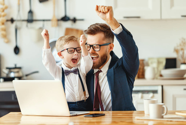 Young male entrepreneur in formal clothes and eyeglasses with little son in formal wear looking at laptop screen and celebrating successful deal while working remotely in modern apartmen - Photo, Image