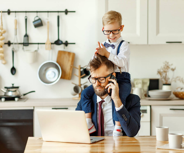 Cheerful playful kid sitting on neck of unhappy busy dad in formal wear during phone conversation and working with laptop in home kitche - Photo, Image