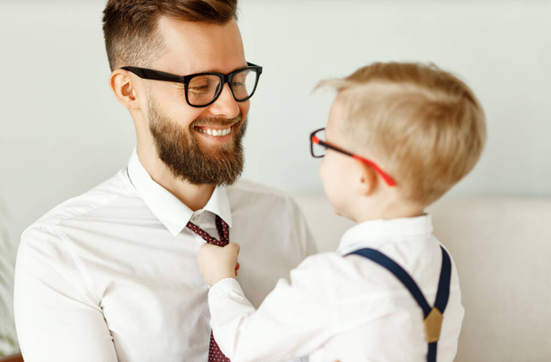 Back view of young child in formal wear and eyeglasses adjusting necktie of cheerful father in similar clothes and glasses while preparing together for even - Foto, imagen