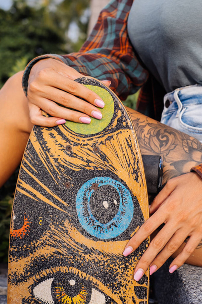 Gros plan de femme tatouée en chemise à carreaux et short en jean avec jolie manucure rose tenir longboard coloré, planche à roulettes - Photo, image
