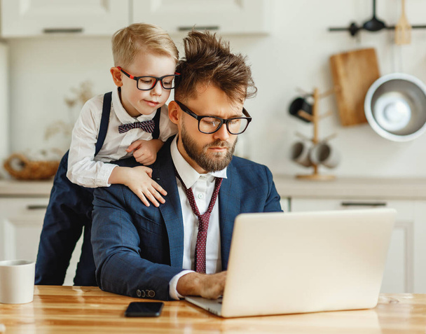Curious little son looking at laptop screen while busy dad in formal outfit with messed up hair working on remote business project at hom - Photo, Image