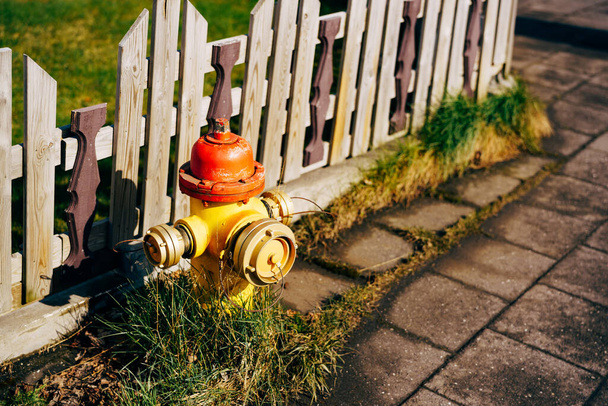 Κίτρινο πυροσβεστικό κρουνό με κόκκινο καπάκι και ασημί καπάκια. Στο γρασίδι κοντά σε ένα ξύλινο φράχτη. - Φωτογραφία, εικόνα