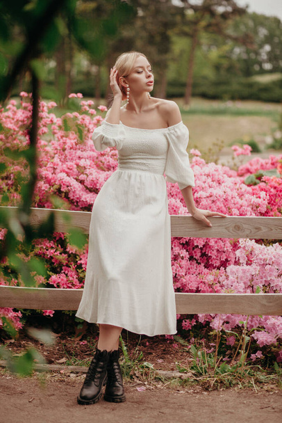 young beautiful woman posing in blooming park. lady in white dress and black boots. pink flowers background. blonde model. romantic spring portrait - Фото, изображение