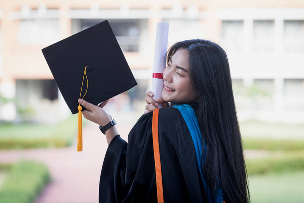 Porträt einer glücklichen und aufgeregten jungen asiatischen Universitätsabsolventin, die am ersten Tag ihres Studiums ein Abschlussgewand und einen Hut trägt und mit einem Abschluss auf dem Universitätscampus feiert. Bildungskonzept. - Foto, Bild