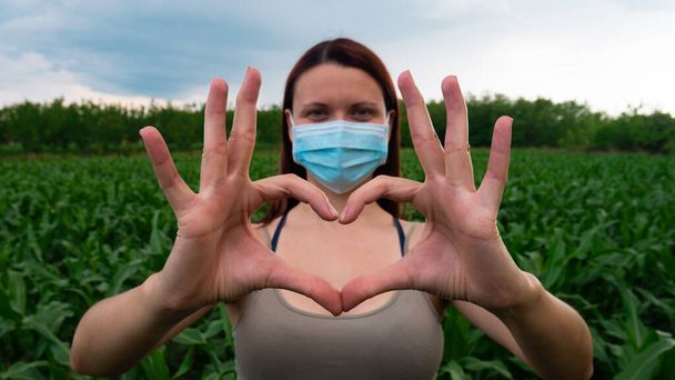 Une femme debout dans un champ de maïs avec un masque médical sur le visage montrant le symbole du cœur avec les mains. - Photo, image