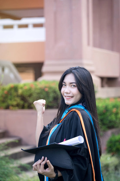 Portret szczęśliwej i podekscytowanej młodej Azjatki absolwentki uniwersytetu nosi suknię dyplomową i kapelusz świętuje z dyplomem w kampusie uniwersyteckim w dniu rozpoczęcia studiów. Koncepcja edukacji. - Zdjęcie, obraz