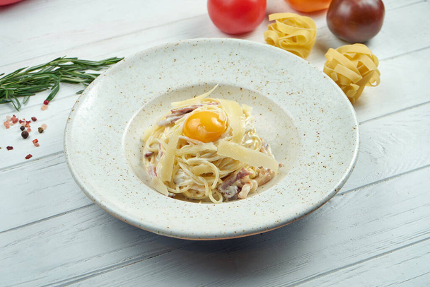 Domowej roboty włoski makaron carbonara z bekonem, parmezanem i żółtkiem w białym talerzu na drewnianym tle. Smaczna kuchnia włoska - Zdjęcie, obraz