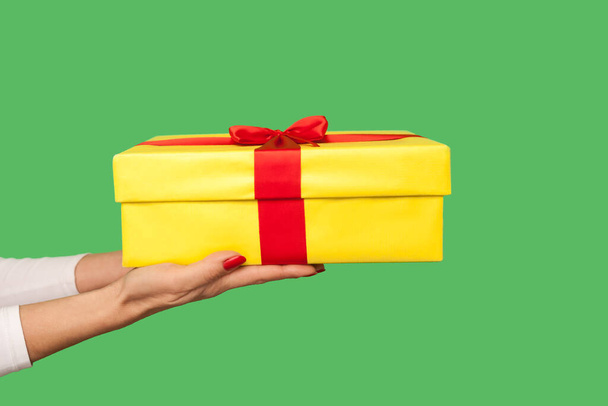 Closeup of woman hand holding gift box with red ribbon, congratulating on birthday, offering present and celebrating Christmas holidays, festive tradition. indoor studio shot isolated on green background - Photo, Image