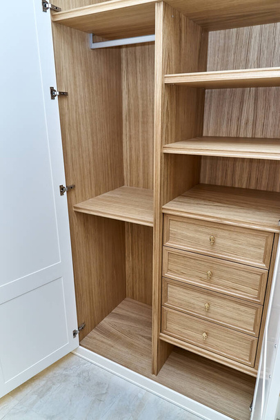 Opened white wardrobe with wooden drawers and shelves. Wooden filling of wardrobe. Wooden wardrobe with white lacquered cabinet doors - Фото, зображення