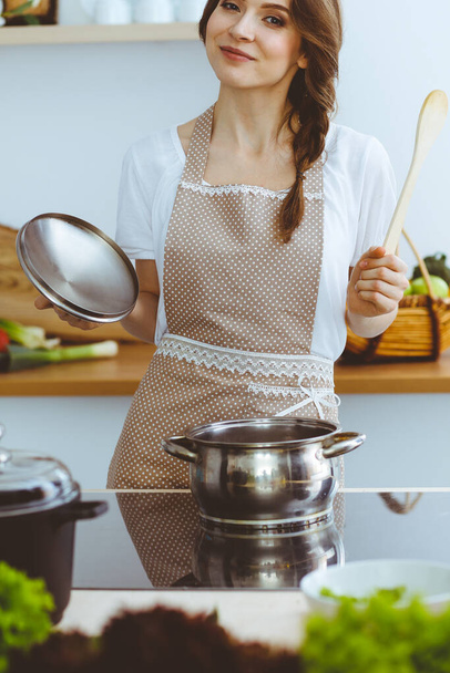 Young brunette woman cooking soup in kitchen. Housewife holding wooden spoon in her hand. Food and health concept - Fotografie, Obrázek