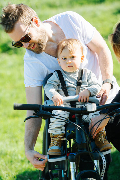 Attentive father carefully strapping his kid to front bike baby chair. Over green countryside. Child looking at camera, he is not very fond of process. - Zdjęcie, obraz